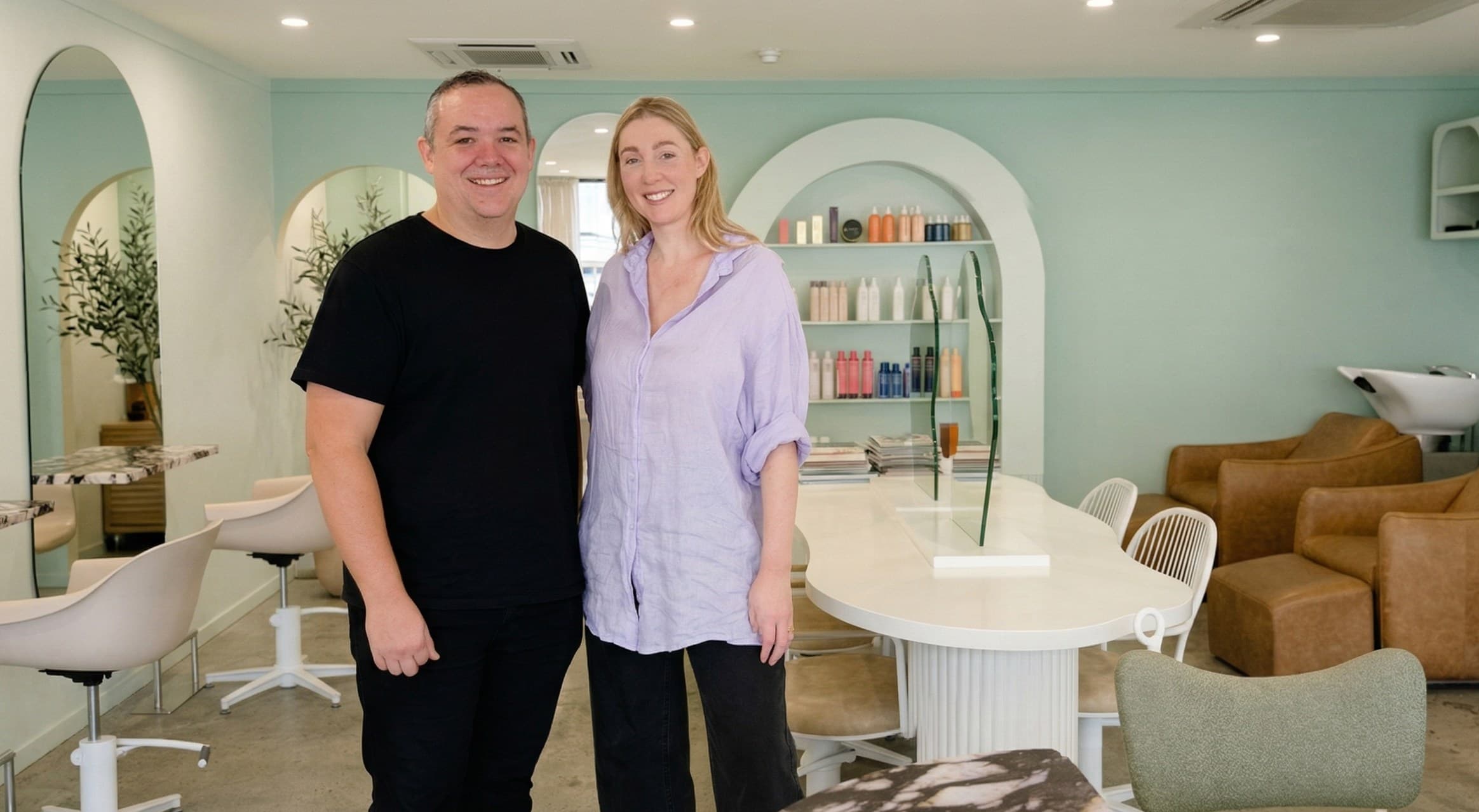 Logan and Jodie, founders of OpenChair, standing together in a modern hair salon with mint green walls, arch mirrors, and leather styling chairs
